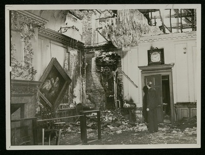 The photograph shows a room devastated by bomb damage. On the left-hand wall, a painting hangs askew. Daylight pours in from a crater in the roof; a chandelier, suspended from the remnants of the ceiling, seems relatively intact. At the corner of the room, a section of the wall has been reduced to rubble, over which half a picture-frame dangles. With his back to us, a man surveys the ruin.